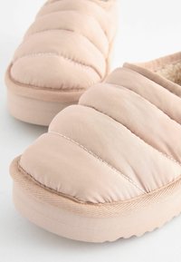 Pantofole slip-on imbottite di un delicato rosa, con un esterno in tessuto liscio, suola texturizzata e dettagli di cucitura sottili. Interno foderato in materiale morbido.