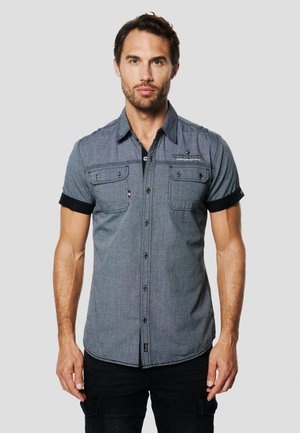 Trumpomis rankovėmis, tamsiai pilko audinio marškiniai su sagomis, dviem krūtinės kišenėmis, juodomis detalėmis ant rankovių ir matomomis siūlėmis.