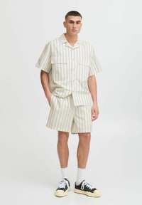 Stripet kortermet skjorte og short sett i kremfarge med tynne mørke striper, med to brystlommer og en avslappet passform, sammen med svarte joggesko.