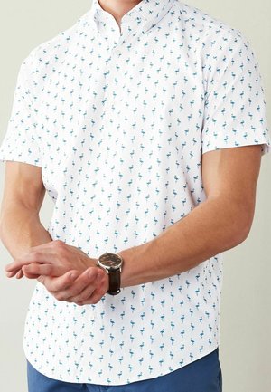 Mężczyzna ubrany w białą koszulę z krótkim rękawem z niebieskim wzorem flaminga, niebieskie spodnie i zegarek na rękę, ręce splecione z przodu.