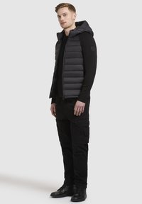 Gilet imbottito nero con cappuccio, caratterizzato da un motivo a cuciture orizzontali, indossato sopra una maglietta nera a maniche lunghe, abbinato a pantaloni cargo neri e stivali.