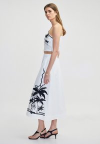 Bílé crop top s černými vyšívanými palmami, spárované s dlouhou bílou sukní vyšívanou černě a černými sandály na podpatku s řemínky.
