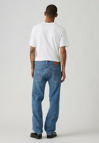 Jeans in denim blu con taglio dritto, design a cinque tasche e una patch in pelle marrone sulla vita posteriore. Abbinati a una maglietta bianca.