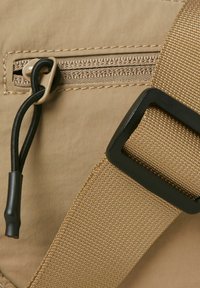 Detail einer tanfarbenen Stofftasche mit einem Reißverschlussfach, schwarzem Zubehör und einem strukturierten Tragegurt, der die Nähte und eine glatte Oberfläche zeigt.