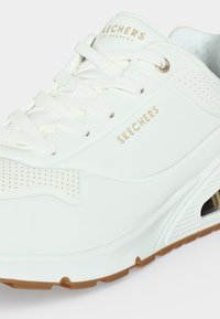 Бяла маратонка Skechers със златно лого, перфорирана странична панел, бели връзки и гумена подметка, видима в долния край.