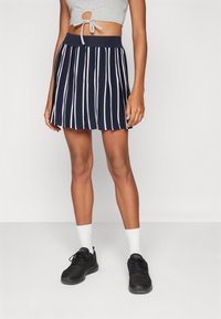 Jupe plissée marine avec des rayures blanches verticales, taille élastique à côtes. Portée avec des baskets noires et des chaussettes blanches de type crew.