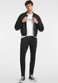 Crna bomber jakna preko bijele grafičke majice, uparena s crnim uskim trapericama i crnim tenisicama s visokim vrhom. Jednostavan, moderan dizajn.