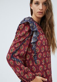 Blusa con motivo paisley in rosso e blu navy, con accenti arricciati blu sulle spalle e maniche lunghe con polsini arricciati.