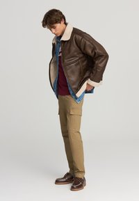 Giacca in pelle marrone con colletto in pelliccia, indossata sopra una camicia di jeans. Abbinata a pantaloni cargo beige e scarpe marroni.