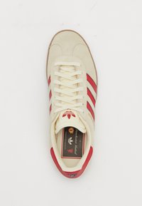 Sapatilha de camurça bege com riscas vermelhas, atacadores crème e sola de borracha. Apresenta o logótipo na língua e na parte traseira, parte superior texturizada e design clássico.