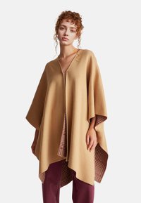 Béžový pončo, s výstřihem do V, vyrobené z měkké tkaniny. Na vnitřní straně má kostkovaný vzor v mahagonové barvě. Volnější střih s širokými rukávy.