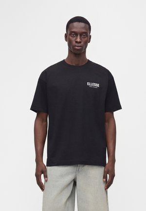 Мъж, облечен в черна тениска с къси ръкави с логото на "ELLESSE" отляво на гърдите и светлосив панталон, стоящ срещу едноцветен фон.