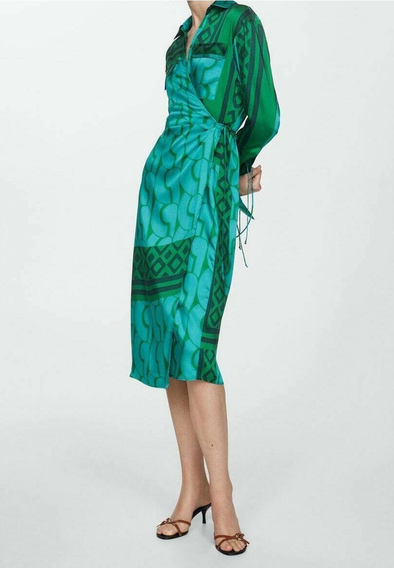 Femme portant une robe portefeuille à motif bleu sarcelle et vert avec des manches longues et des sandales marron à talons et à lanières, debout devant un fond uni.