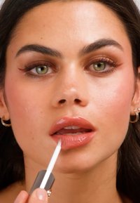 Jonge vrouw met groene ogen die roze lipgloss aanbrengt, met gouden oorringen en vleugelvormige eyeliners.