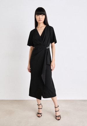 Black wrap dress with short sleeves, V-neck, and a tied side accent. Smooth fabric, mid-calf length, paired with strapped black sandals.
