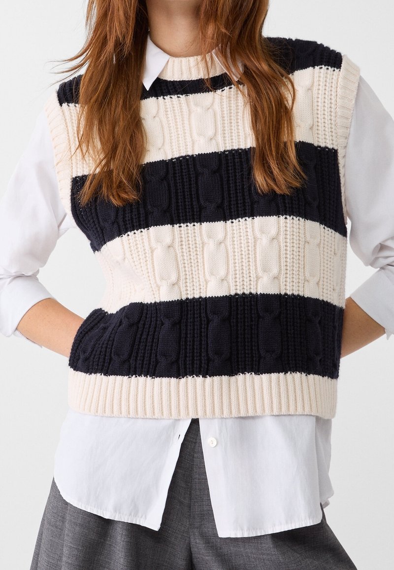 Femme portant un pull sans manches en maille torsadée rayé noir et blanc par-dessus une chemise blanche boutonnée avec un pantalon gris.