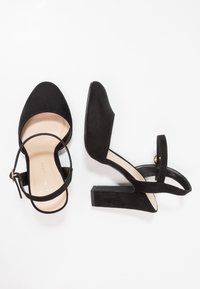 Černé semišové boty na vysokém podpatku s otevřenou zadní částí, kotníkovým páskem a mohutným podpatkem. Podešev je béžová a má hladkou texturu.