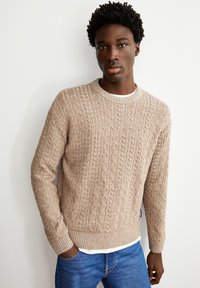 Sweatshirt en tricot beige avec un col rond, des poignets et un ourlet côtelés. Associé à un jean bleu, mettant en valeur un tissu texturé.