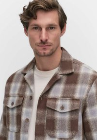 Brown and blue plaid jacket with a buttoned front and chest pockets, crafted from a textured fabric, layered over a light-colored shirt.