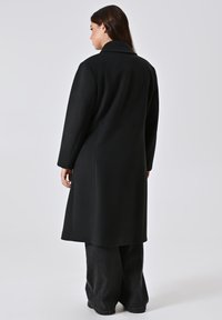 Cappotto nero lungo a doppio petto con colletto a revers, tessuto liscio, tasche laterali e vestibilità sartoriale, abbinato a jeans neri a gamba larga.