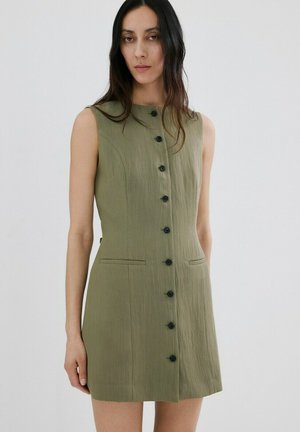 Mujer con vestido sin mangas verde oliva abotonado con bolsillos de ojal delanteros, de pie contra un fondo claro y liso.