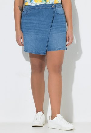 Person trägt einen blauen Jeans Wickelrock, weiße Turnschuhe, ein geblümtes Oberteil und ein Tattoo am linken Unterarm, stehend vor weißem Hintergrund.