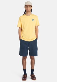 T-shirt in cotone giallo con logo verde sul petto, abbinata a pantaloni corti blu navy con due tasche frontali e calzature marroni.