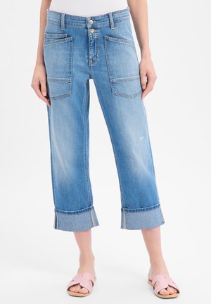 Jean bleu clair à jambes larges et raccourci, avec grandes poches plaquées à l'avant et revers roulés, porté avec des sandales plates ouvertes rose.