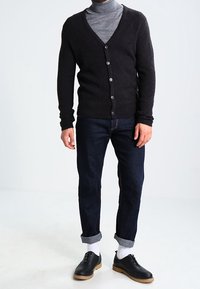 Cardigan di lana grigio scuro con scollo a V e chiusura a bottoni, indossato sopra un dolcevita grigio chiaro, abbinato a jeans scuri e scarpe nere.