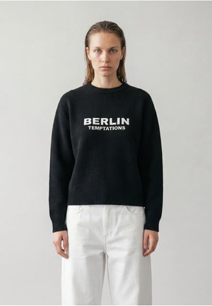 Un mannequin est debout devant un fond gris clair, portant un pull noir "Berlin Temptations" et un pantalon blanc.
