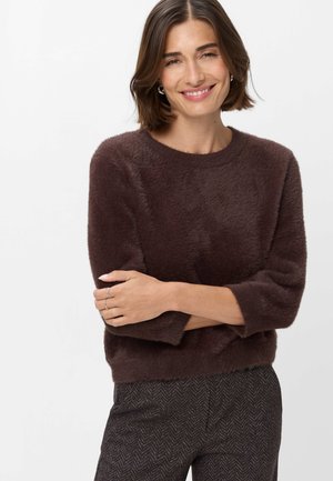 Kvinde med kort brunt hår iført en blød mørkebrun sweater og mønstrede mørke bukser, smilende med armene krydset på en hvid baggrund.