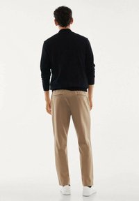 Uomo con maglietta nera a manica lunga, pantaloni beige e sneakers bianche, in piedi di spalle su uno sfondo chiaro e uniforme.