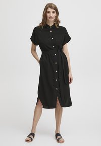 Rochie neagră, cu nasturi, cu mâneci scurte, talie cu legătură și fante laterale, realizată dintr-un material texturat. Are un buzunar pe piept și nasturi aurii.