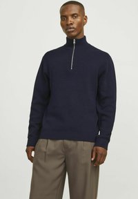 Pulover cu guler cu fermoar de culoare bleumarin dintr-un material texturat. Are mâneci lungi și manșete ribbate. Purtați-l cu pantaloni pliți de culoare maro deschis.