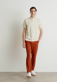 Beige polo skjorte med en to-knapps placket, parret med rustfargede bukser. Fottøy er hvite sneakers med lys såle. Minimalistisk setting.