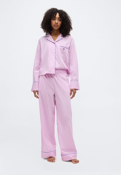 Femme debout pieds nus, portant un pyjama à manches longues rayé rose et blanc avec un biais bleu sur les poignets et le col.