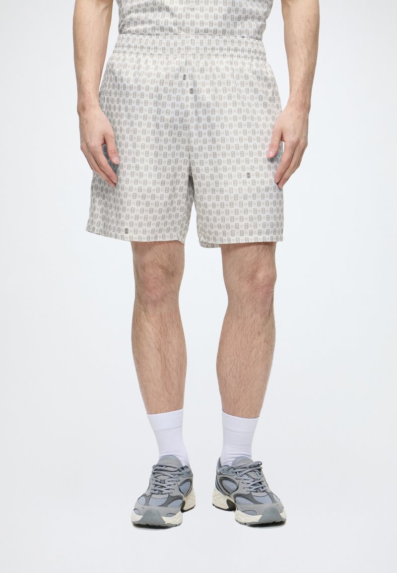 Homme portant un short gris clair à motifs, des chaussettes blanches et des baskets grises, debout devant un fond uni.