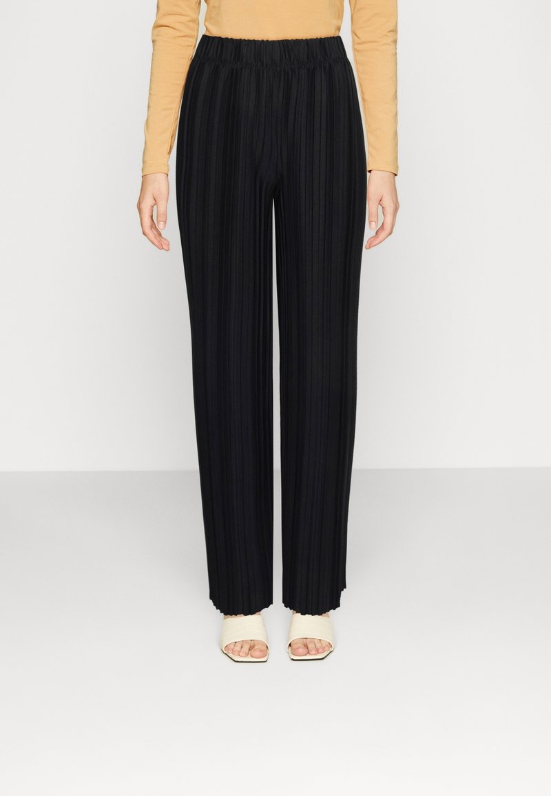 Kvinna iförd svarta plisserade vida byxor, beige långärmad topp och vita öppna klacksandaler, som står framför en enfärgad bakgrund.