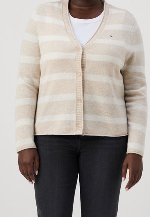 Mørkhudet person iført en beige og hvide stribet cardigan over en hvid top og sorte bukser, stående med armene afslappet ned langs siden.