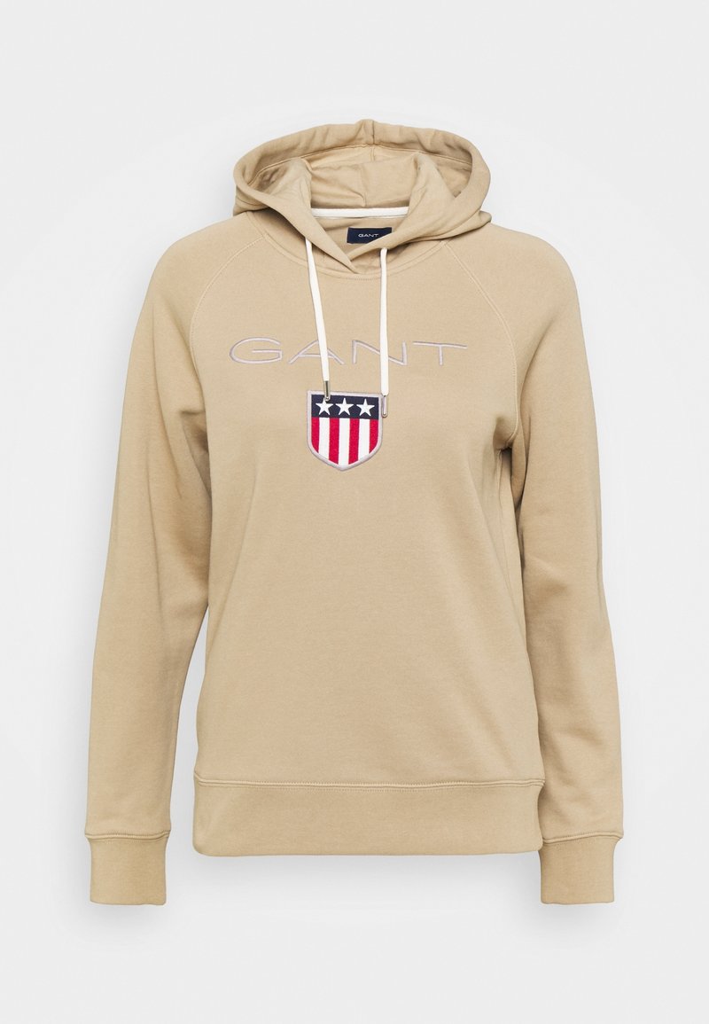 Sweat à capuche beige avec cordons blancs, texte "GANT" et écusson en forme de bouclier comportant trois étoiles blanches au-dessus de bandes rouges et blanches sur la poitrine.