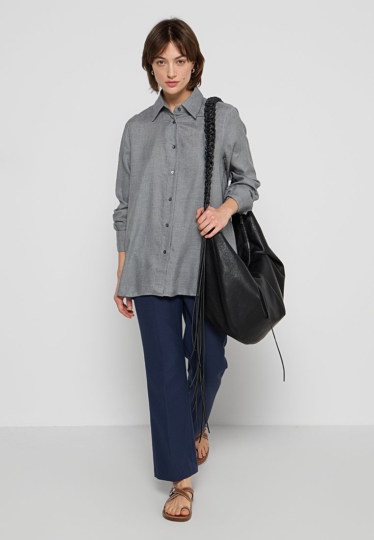 Graues Hemd mit langen Ärmeln, kombiniert mit marineblauen, weit geschnittenen Hosen und braunen Sandalen. Eine schwarze Lederhandtasche mit geflochtenem Tragegurt wird gehalten.