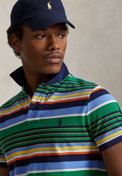 Ung mann som ser til siden, iført en mørk blå caps og en flerfarget stripete poloskjorte med hevet krage.