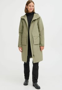 Olivengrønn vanntett parkas med hette, to store frontlommer og glidelås. Brukt over svart undertøyslag og bukser.