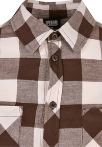 Camicia a maniche lunghe a quadri marrone e crema con colletto, dotata di due bottoni marroni e di una tasca sul petto sul lato sinistro.
