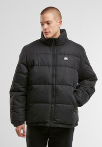 Sort pufferjakke med høj krave, quiltet design, lynlås-lukning og et lille brandlogo på brystet. Glat, skinnende stoftekstur.