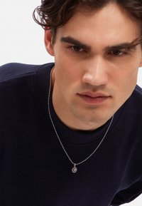 Silver necklace with a circular pendant featuring a geometric design, worn over a textured navy blue shirt. Close-up of the wearer’s upper body.