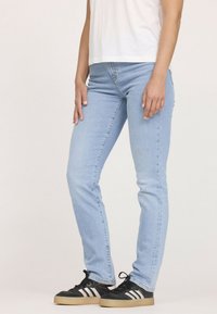 Jean en denim bleu clair au fit slim, avec des coutures contrastantes et un design classique à cinq poches, porté avec des chaussures de sport noires.