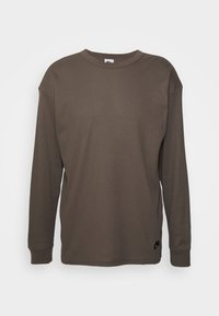 Hnědá košile s dlouhým rukávem z texturované látky. Design s kulatým výstřihem, pohodlný střih a žebrované manžety. Na lemu je malá černá logo.