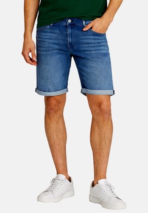 Mann trägt hochgekrempelte blaue Denimshorts, weiße Sneakers und ein grünes Hemd, steht mit einer Hand in der Tasche.