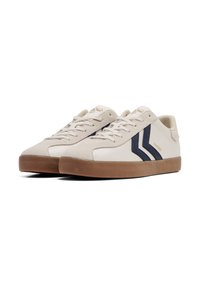 Tenisi din suede bej cu accente în dungi bleumarin, talpă din cauciuc și șireturi plate. Prezintă laterale texturate și o etichetă cu marcă pe limbă.